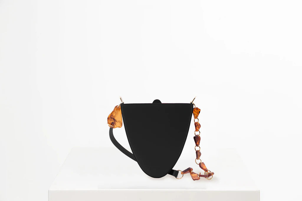 Flap topped oval structured black calf leather cross body bag with an adjustable leather and natural resin chain linked strap on a white stand
