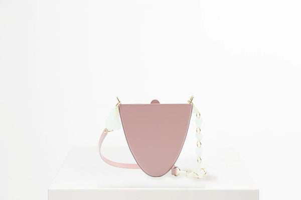 Flap topped oval structured dusty rose calf leather cross body bag with an adjustable leather and natural resin chain linked strap on a white stand