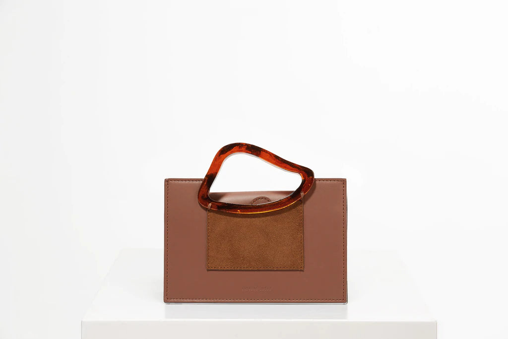 Rectangle shaped chocolate brown calf leather saddle bag with natural resin handle holder and remowable calf leather handle strap on a white stand