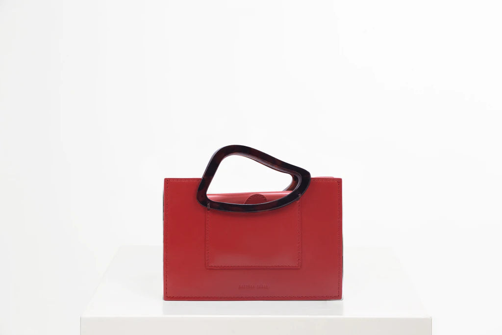 Rectangle shaped red calf leather saddle bag with natural resin handle holder and remowable calf leather handle strap on a white stand