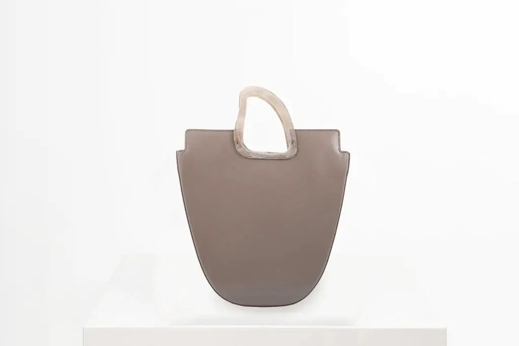 Flat topped oval structured taupe calf leather tote bag with natural resin round handles and a removable adjustable calf leather strap on a white stand