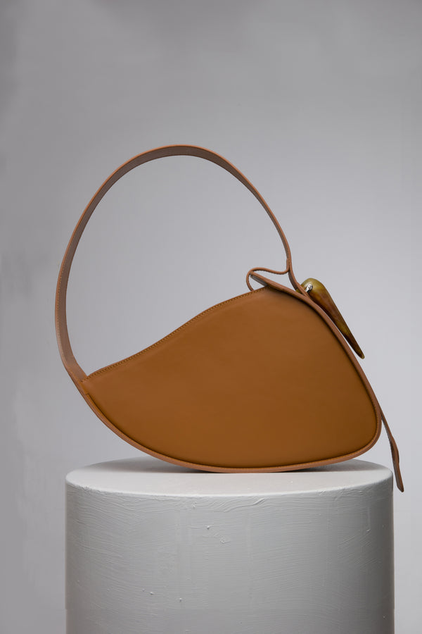 Flat topped oval structured saddle bag with bio resin accessories and non-removable but adjustable calf leather strap on a white stand