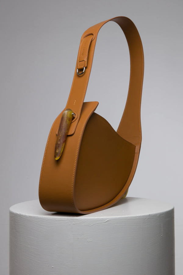 Flat topped oval structured saddle bag with bio resin accessories and non-removable but adjustable calf leather strap on a white stand